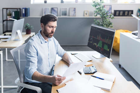Confident business man stocks broker sitting in front of monitor screen in office analyzing stat of index charts data analyzes market trend. Forex trading agency worker agent reviewing profits growthの写真素材