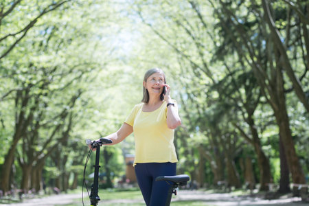 Senior happy woman on a walk in the summer park, smiling and chatting on the phone with a bicycle