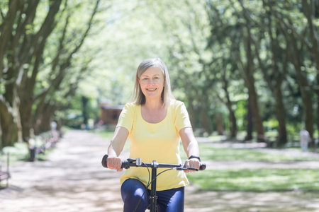 Active retired woman rides a bicycle in a summer park, a senior gray-haired retired woman walks