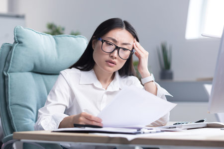 Young Asian woman working on paper in office, businesswoman anxiously examines documents and...