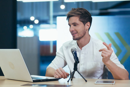 Interview recording. Young handsome man sitting at a table in front of a microphone and laptop,...