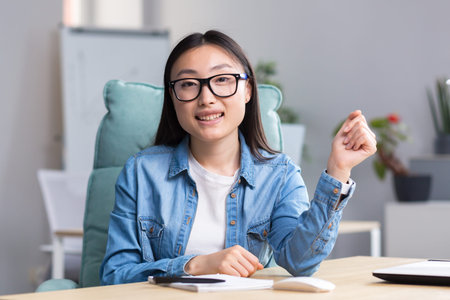 Young beautiful Asian woman talking on video call, business woman working in modern office, smiling...
