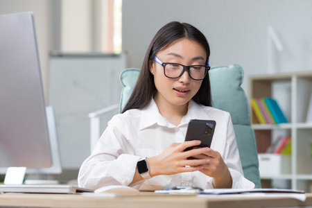 Young beautiful Asian business woman works in modern office, uses phone, smiles, reads good news, chats in app with friendsの写真素材