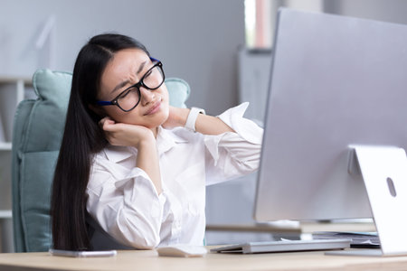Overtired Asian woman office worker, has severe neck pain, massages neck muscles with hand, female...