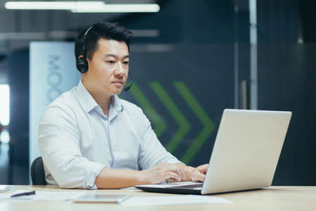 Successful asian employee boss businessman working with laptop using headset for video call, man at work in officeの写真素材