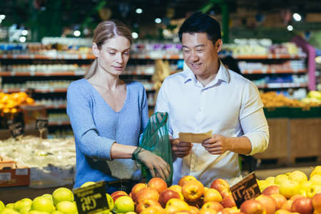 happy mixed race asian couple family man and woman choosing fruits or vegetables in grocery store supermarket. Joint daily shopping together. Buyers customer select product pick an apple in eco bagの写真素材
