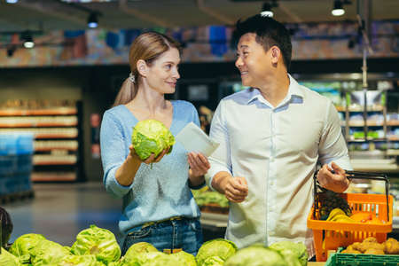 Happy mixed race asian couple family man and woman choosing vegetables in grocery store supermarket. make a purchase daily shopping. Customer select product pick an cabbage in eco bag in food marketの写真素材