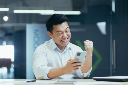 Success win. Young Asian business man holds phone in hand, rejoices win, good news, reads message....