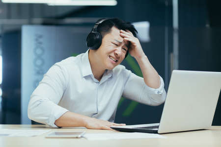 Asian businessman watching sports match in office, man looking disappointed in laptop, using headphones to listenの写真素材