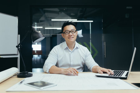 Portrait of successful asian male architect smiling and looking at camera, working on project plan drawing in design studio officeの写真素材