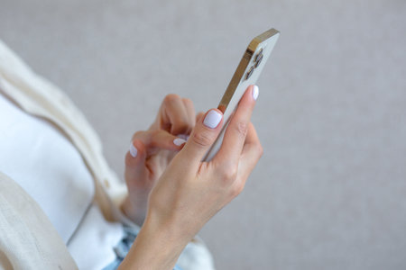 A close-up photo of a beautiful womans hand with a white manicure is using a gold-colored phone,...