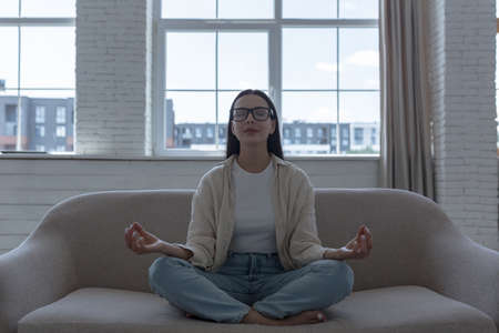 Woman alone at home meditating in front of window sitting on sofa, beautiful girl calm down and resting in lotus pose, wearing glasses with closed eyesの写真素材