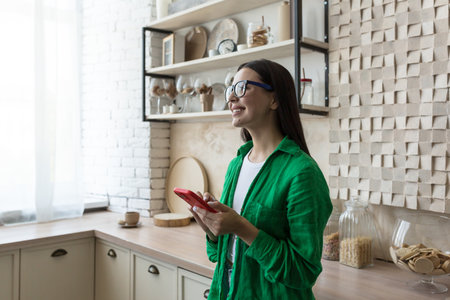 Young beautiful woman in glasses and green shirt near the window at home in the kitchen, using a red phone, smiling and looking dreamily in the windowの写真素材