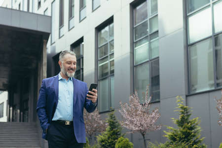 Senior experienced gray-haired businessman outside office, smiling and happy using phone, boss reading and looking at smartphone screen, walking near buildingの写真素材