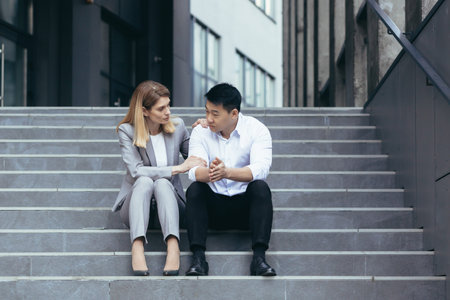 Woman taking care of disappointed asian businessman with failed business sitting on stairs in city center, friendly support from co-workerの写真素材