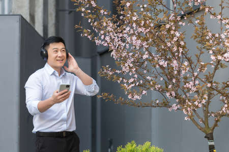 Cheerful and smiling successful Asian business man listening to music using smartphone and big headphones, manager resting outside office in shirtの写真素材