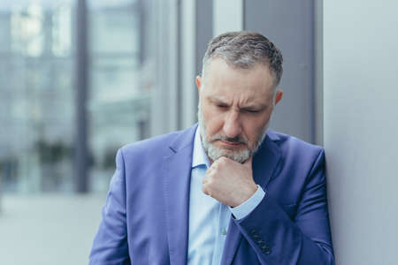 Senior gray haired man outside office building, standing and thinking, confused and sad businessman boss in business suit, over decisionの写真素材