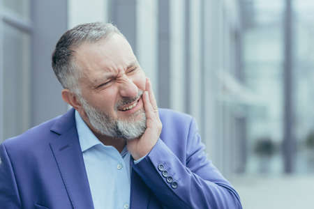 Close-up photo portrait of senior gray-haired businessman, experienced businessman outside office building, having severe toothacheの写真素材