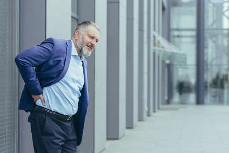 Overtired senior gray-haired man outside office building, businessman in business suit, holding hand behind back, having severe back pain, long sedentary workの写真素材