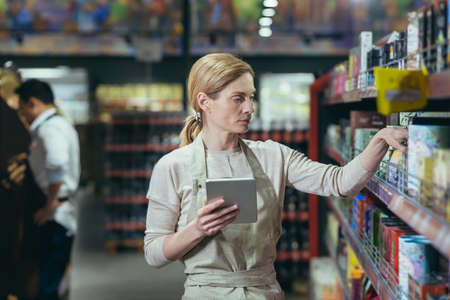 A woman seller in a supermarket uses a tablet computer to count the remaining goods, colleagues conduct an inventory in the grocery department among the shelves with goodsの写真素材