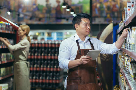 Two workers in a supermarket in the grocery department arrange products, an Asian man with a tablet computer and a woman in aprons among the products on the shelves and racksの写真素材