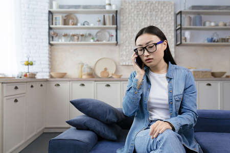 Upset young beautiful Asian woman in glasses talking on the phone sitting on the sofa in the living roomの写真素材