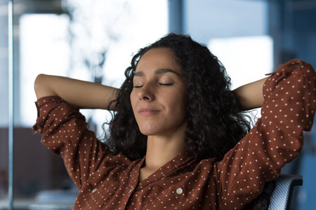 Close-up photo portrait of young beautiful curly Arab woman resting indoors at home, woman with hands behind head resting sitting on sofa near window, eyes closed breathing, meditating and dreamingの写真素材
