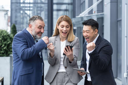 group of colleagues employees read good news looking at smartphone. Happy team rejoices about successful investment while using, browsing mobile phone. Standing outside near office building.の写真素材