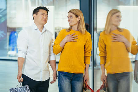 multiracial couple asian man and caucasian woman walking together in mall after shopping with colored bags in hands. walking past shop windows with clothes cheerful family hugging smile.の写真素材