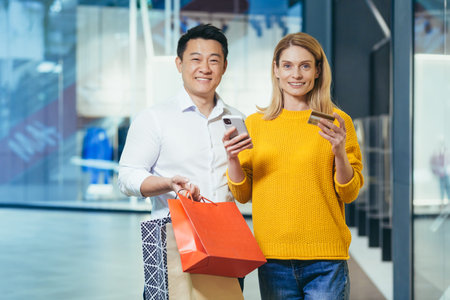 Portrait of happy shoppers asian man and blonde woman smiling and looking at camera, diverse couple in supermarket with packages and colorful shopping bags holding smartphone and bank credit cardの写真素材