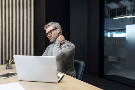 Senior mature businessman working inside office building, gray haired man sick and tired massaging neck with hand, boss having severe pain in neck and back.の写真素材