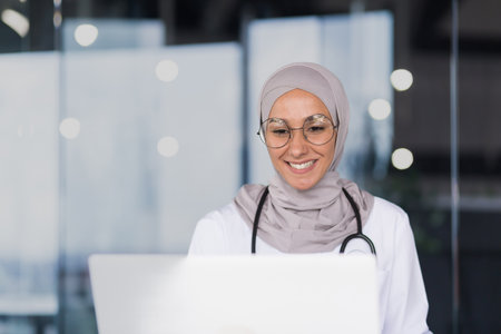 Close-up photo portrait of a female doctor in a hijab, a Muslim woman works in the office of a modern clinic, a female doctor in a gown and glasses uses a laptop.の写真素材
