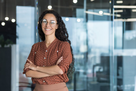 Young beautiful and successful hispanic businesswoman smiling and looking at camera, female worker working inside office wearing glasses and curly hair, arms crossed portraitの写真素材