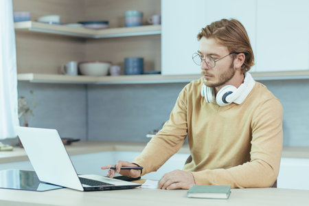 Young serious and focused man studying remotely at home, student in kitchen wearing glasses using...