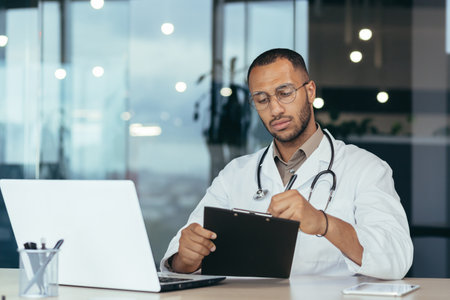 Serious and focused hispanic doctor in medical coat and stethoscope behind paperwork, man in glasses working inside clinic office using laptop.の写真素材