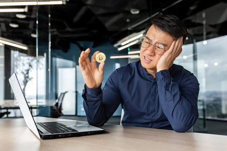 Frustrated asian businessman investor holding crypto currency bitcoin in hands, man working inside...