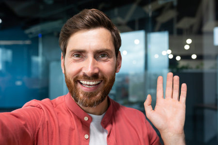 Young happy businessman looking into smartphone camera and talking on video call, man working inside...
