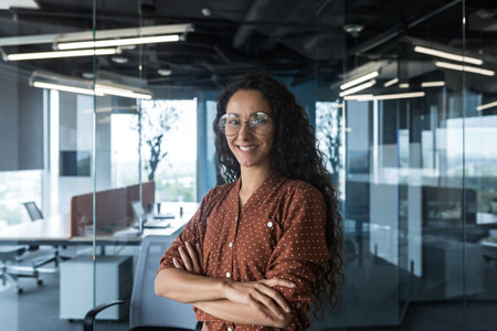 Portrait of successful young beautiful woman company owner and founder. Hispanic businesswoman standing in office wearing glasses and brown shirt, looking at camera, arms crossed, smiling.の写真素材