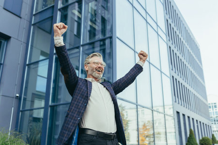 Successful and happy gray-haired boss outside office building rejoices success and victory, triumphs with hands up, businessman business owner in business suit and glasses, mature investor banker.の写真素材