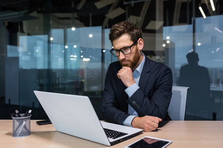 Serious thinking businessman working inside office building, man pondering important decision, boss in business suit using laptop at work.の写真素材