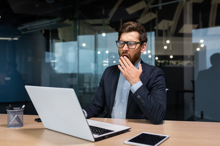 Mature bored businessman working inside modern office building, boss in glasses and business suit using laptop, investor yawning sitting on chair at table.の写真素材