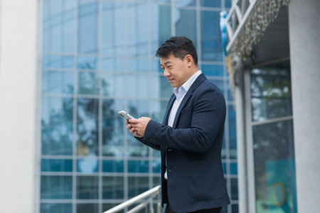 Successful Asian businessman in business suit walking outside near office building, male manager director using phone, smiling and happy reading online message.の写真素材