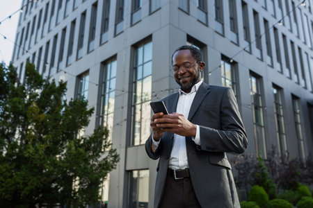 Mature african american boss using phone, businessman outside office building in business suit and glasses, senior man typing message and browsing online pages smiling.の写真素材