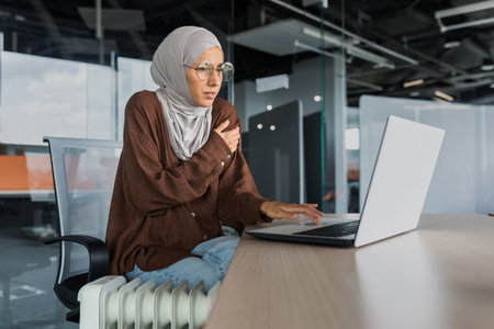 Businesswoman freezing in office at work, woman in hijab near heater trying to keep warm working inside office building with laptop, cold in office.の写真素材