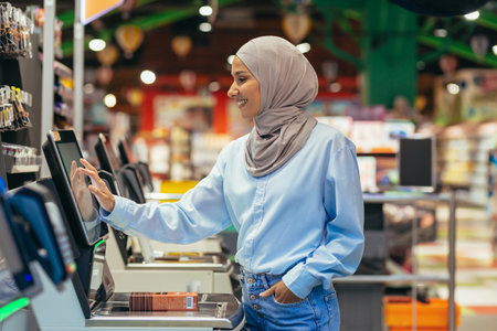 A woman buyer in a supermarket in a hijab pays for goods at a self-service checkout, convenient service for customers.の写真素材