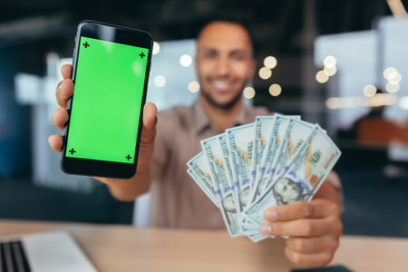 Selective focus, businessman working inside modern office building, african american man with laptop looking at camera showing smarphone ekras and winning dollars money cash.の写真素材