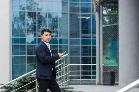A young handsome Asian man goes to a meeting, an interview for a job in an office center. He is standing on the stairs with a phone and documents in his hands, looking at the camera.の写真素材