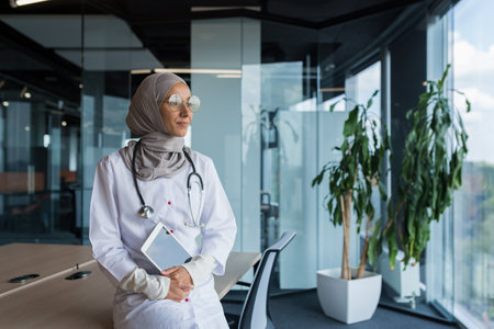 A happy and thoughtful young Muslim woman, an Arab doctor in a hospital, is sitting at her desk, holding a tablet in her hands, looking out the window, smiling, loving her work.の写真素材