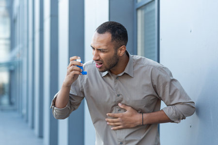 An attack of asthma, allergies. A young Hispanic, African American man is standing outside using an inhaler, experiencing chest pain and shortness of breath. Needs medical help.の写真素材