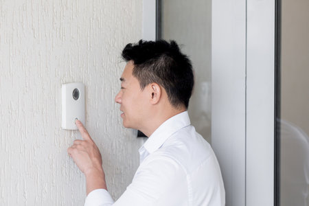 Close-up photo. A young Asian man calls with his finger on the electronic doorbell on the wall of the house, office. Speaks into the intercom.の写真素材